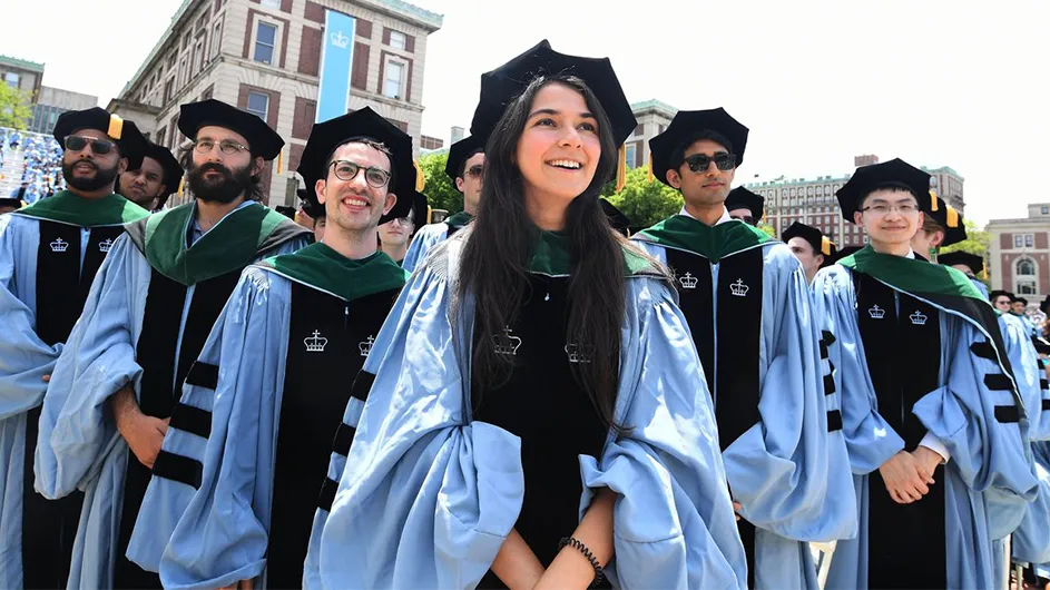 Columbia doctoral graduates stand in blue gown, tam, and green hood. 