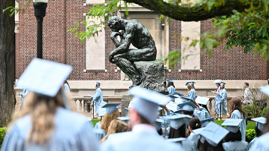 Grads in blue cap and gown circle the thinker statue on Columbia University's Morningside Heights campus.