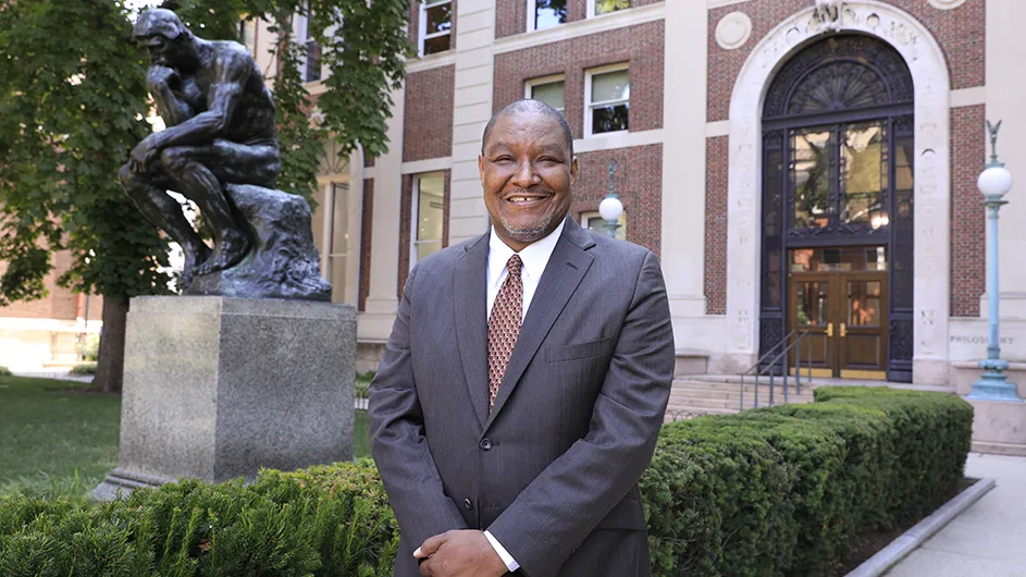 A man in a grey suit, standing outside in front of a brick building and a statue of a man sitting, thinking, resting his head on his hand.